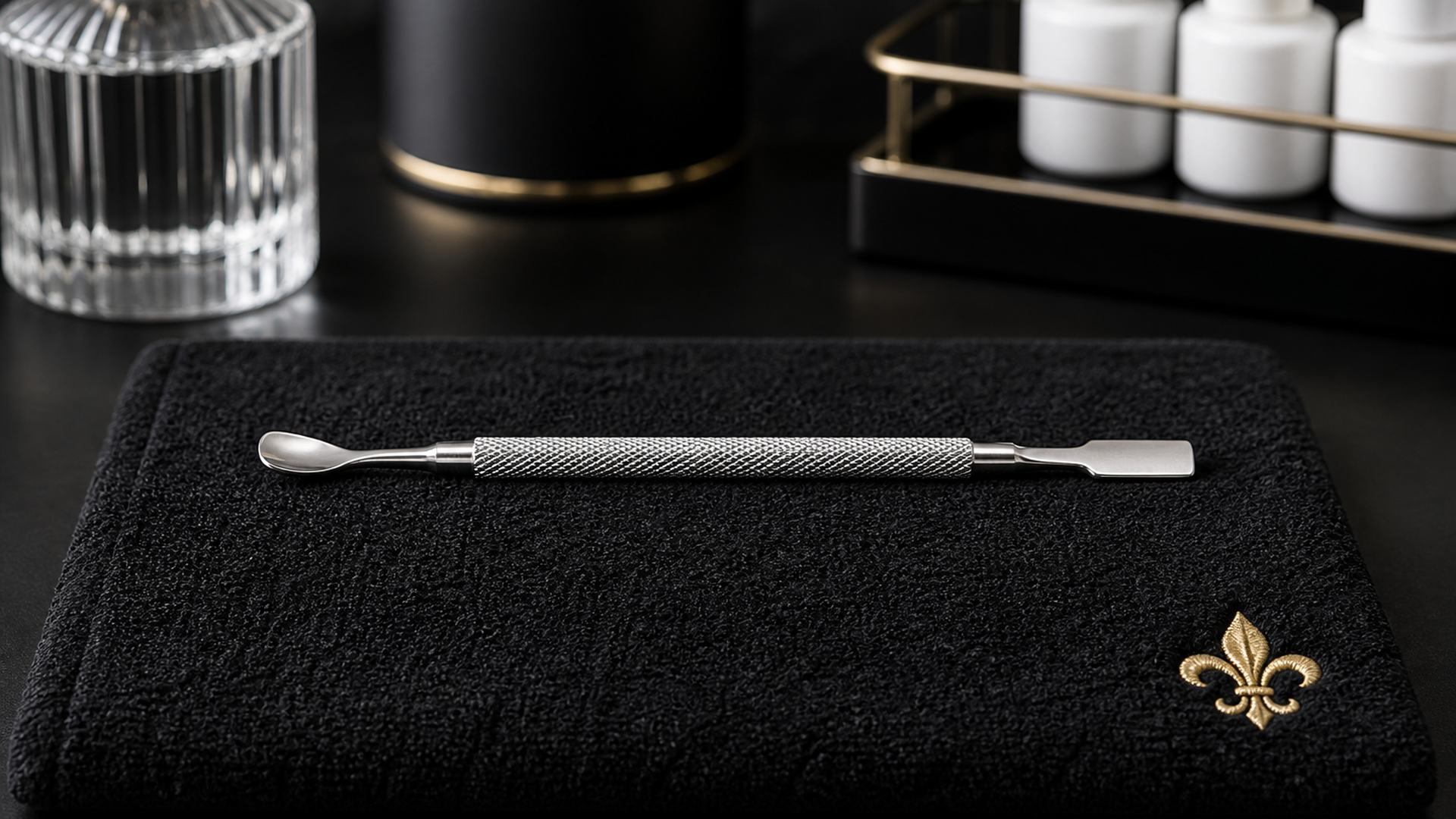 Single stainless dual-ended cuticle pusher on a matte black manicure station with brushed gold fleur accent — Lou Essentials